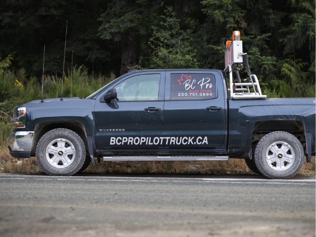 A single BC Pro Pilot Truck, equipped with an oversized load escort sign, stands parked along a forested roadside. The truck, marked with the 'bcpropilottruck.ca' logo and contact information.