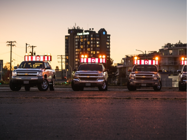 As dusk sets in, a trio of BC Pro Pilot Trucks with illuminated 'D' signs are lined up on the urban roadway, showcasing their readiness for nighttime escort operations for oversized loads.